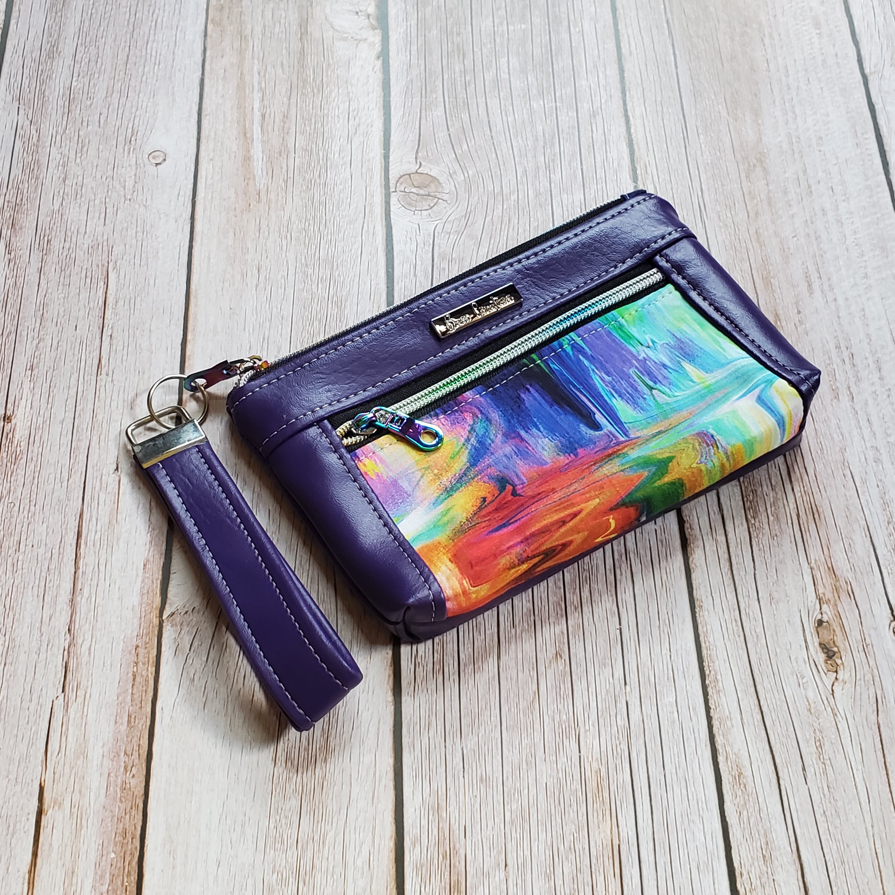 Side view of of a medium makeup pouch with a colorful blurred print center panel and purple faux leather accents and a silver metal Bass Creations logo.