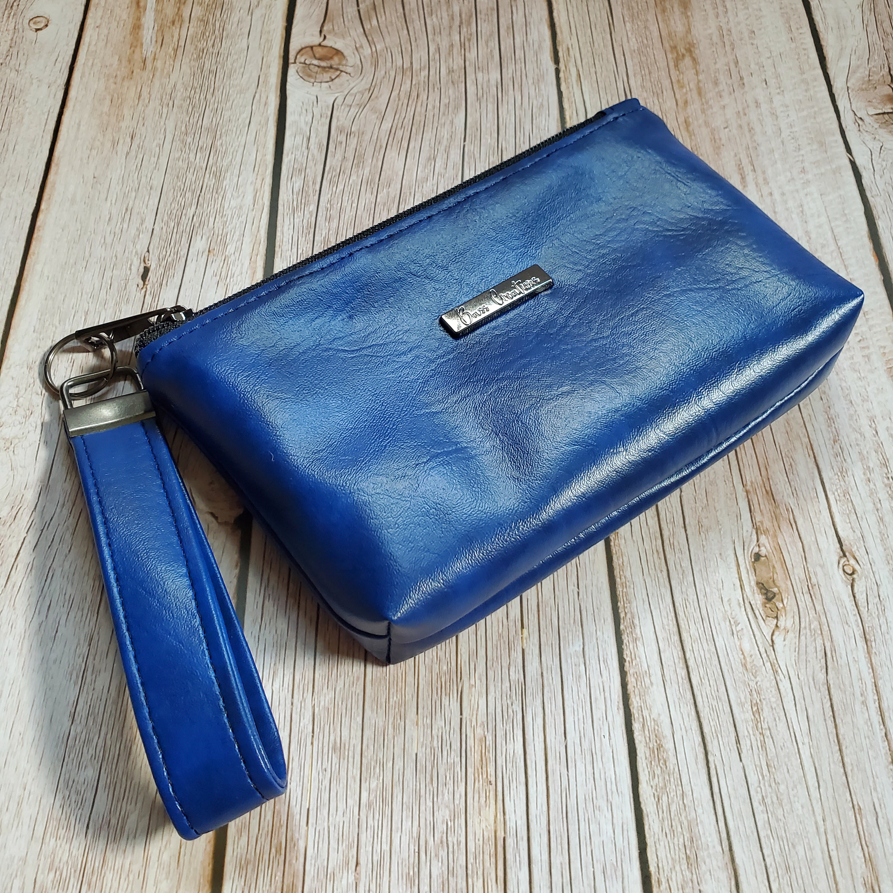 side view of a blue marbled faux leather makeup bag with gunmetal hardware and a gunmetal Bass Creations logo. It also includes a wristlet strap for easy carrying.