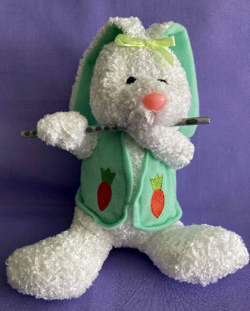 Plush Easter Bunny Flutist made of white chenille and wearing a green vest decorated with carrots, black bead eyes, chiffon yellow hair bow, and pink jelly bean like nose. SO CUTE. Playing a silver wood flute. An ideal basket stuffer for the flutist in your life. She is a sitter for easiness of placement.