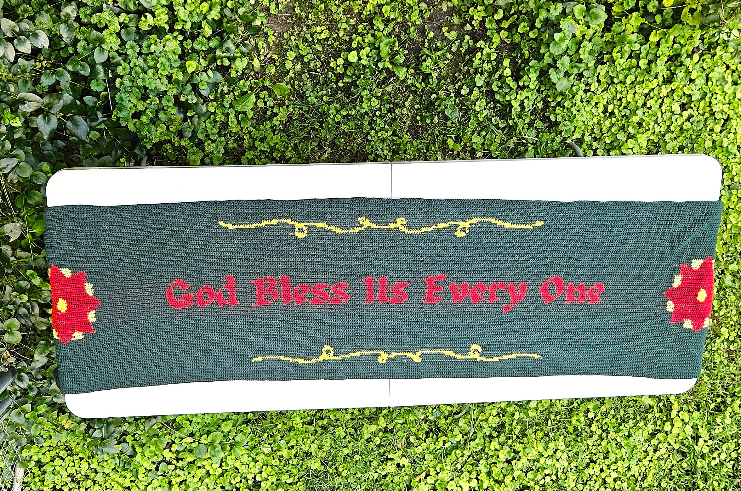 A dark green and red table runner with God Bless Us Every One in the center, poinsettias on either end. And yellow swirls around the quote. A dark green and red table runner with God Bless Us Every One in the center, poinsettias on either end. And yellow swirls around the quote.