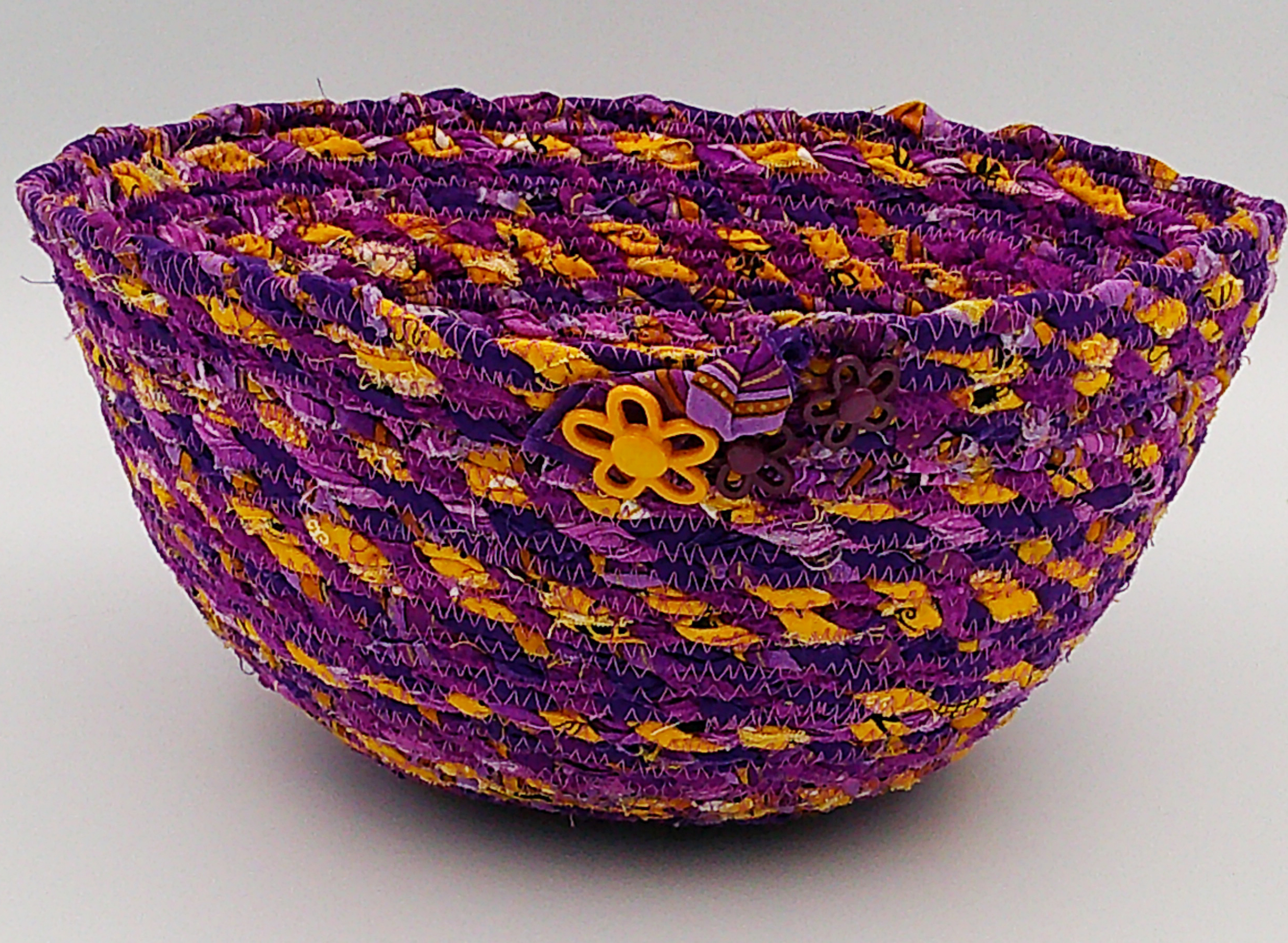 This shows the detail of the front of the basket. There is one large golden yellow flower button with 2 smaller purple ones. This shows the detail of the front of the basket. There is one large golden yellow flower button with 2 smaller purple ones.