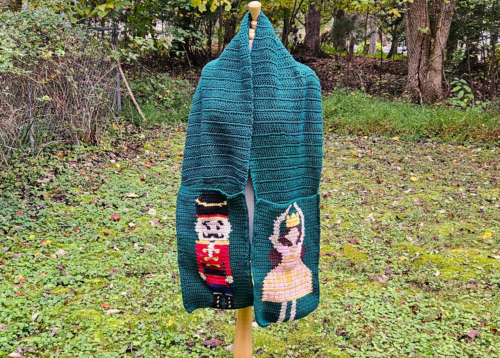 A green crochet pocket scarf on a dress form. There is a nutcracker on the left pocket, and a ballerina in a pink dress on the right pocket. A green crochet pocket scarf on a dress form. There is a nutcracker on the left pocket, and a ballerina in a pink dress on the right pocket.