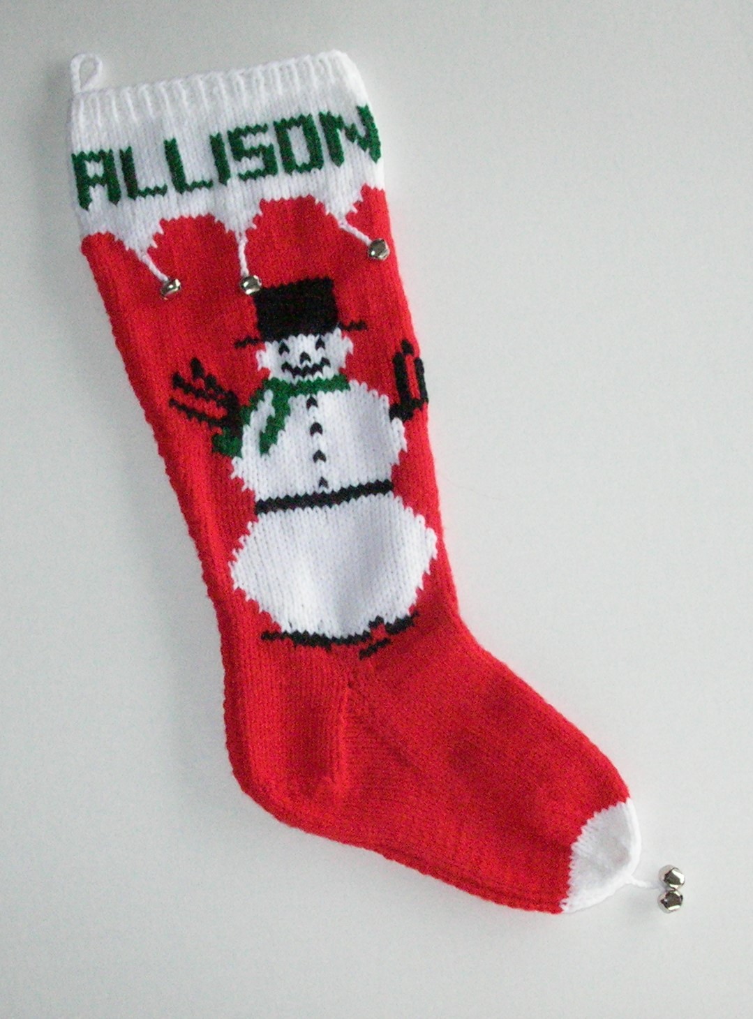 Snowman with green scarf on red and white hand knit Christmas stocking, personalized at top, and embellished with silver bells.