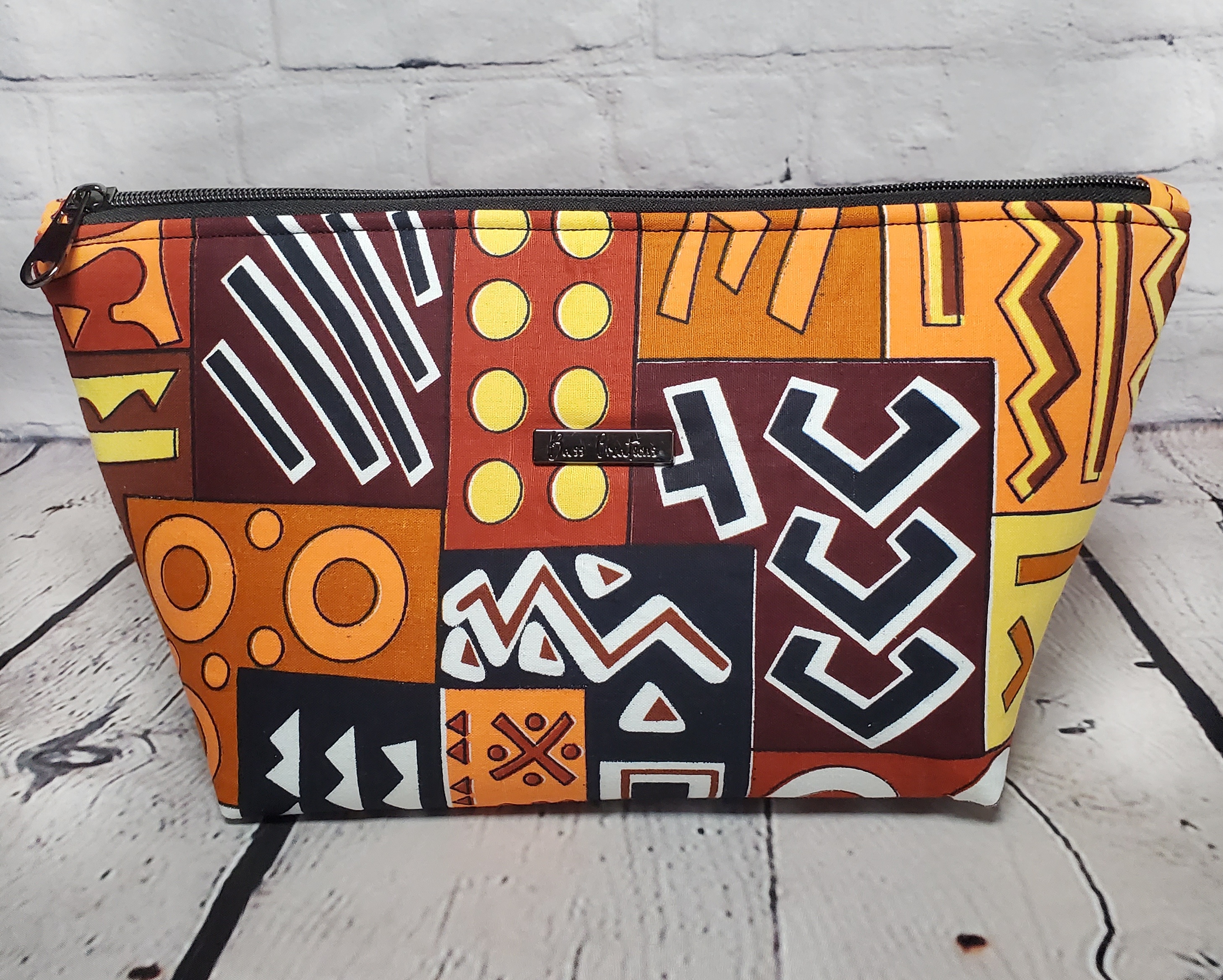 Front of a brown orange and yellow geometric print cosmetic bag with a gunmetal zipper and Bass Creations logo.