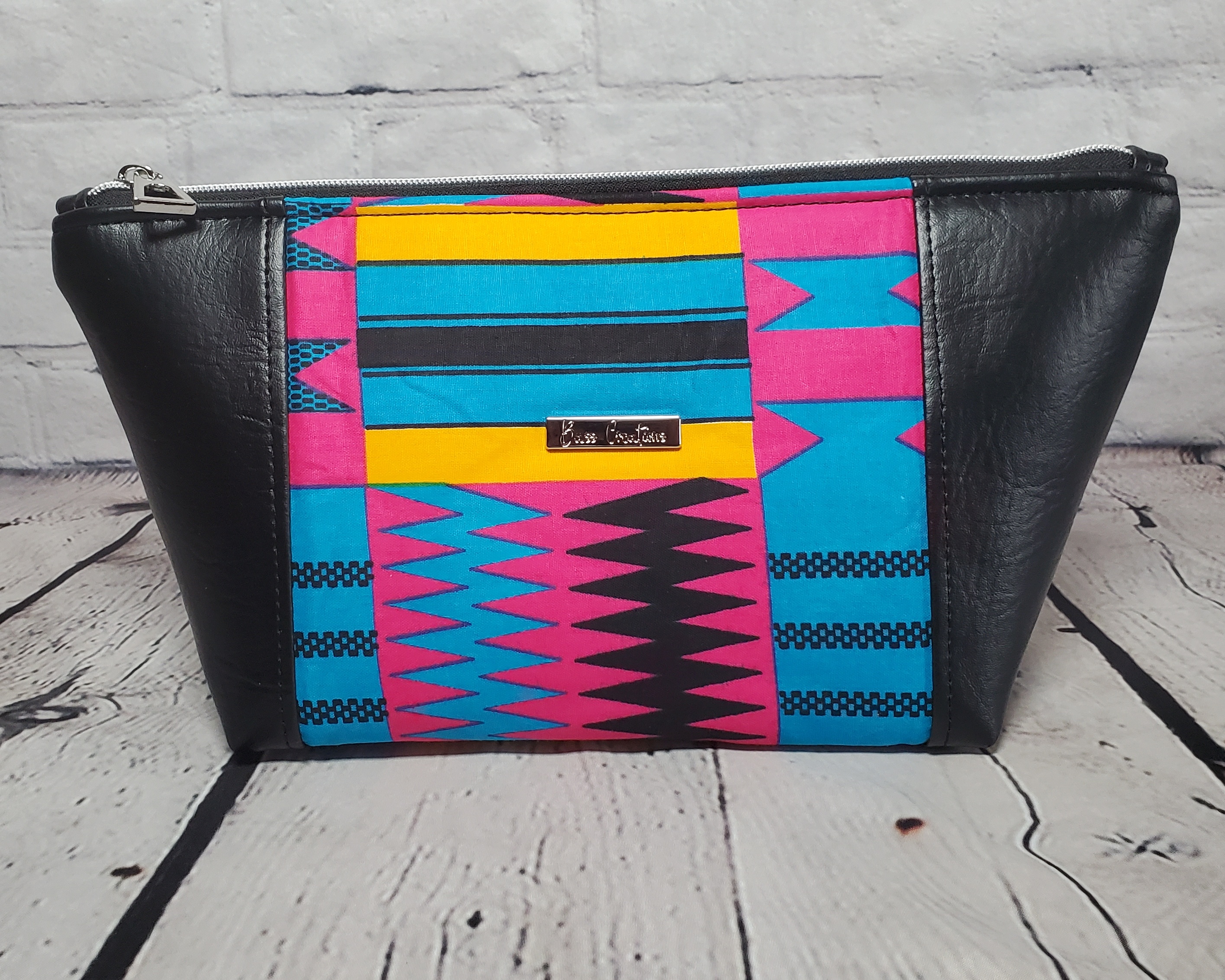 Pink blue and orange geometric print makeup bag with black faux leather accents, silver top zipper and silver Bass Creations logo.